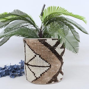 Puede incluir: Una maceta tejida con un patrón geométrico en crema, marrón y marrón oscuro contiene una planta verde con hojas largas y delgadas. Una guirnalda floral azul descansa cerca de la maceta. La maceta es cilíndrica y tiene una apariencia texturizada.