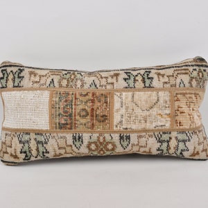 May include: A rectangular cushion with a patchwork design. The cushion has a beige background with rectangular patches in beige, brown, and green. The edges are trimmed with a darker beige border. This cushion is likely for home decor and measures approximately 40cm in length.