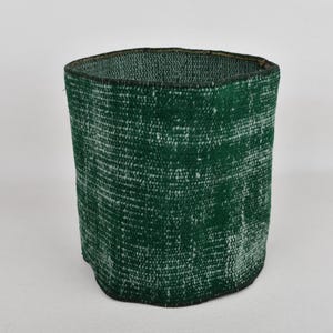 May include: A cylindrical, open-top container with a dark green and white textured pattern. The container has a slightly irregular shape and a dark rim. The item appears to be made of a woven material, possibly fabric or rug material.