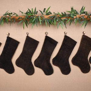 May include: A row of seven dark brown Christmas stockings hanging from small hooks. A bamboo branch with green leaves is above the stockings. The stockings are made of a textured material.