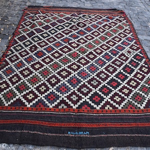 Puede incluir: Una alfombra turca tradicional con un patrón geométrico en tonos de rojo, azul, blanco y negro. La alfombra tiene un borde marrón oscuro con una inscripción tejida que dice "#HALIIBRAM".