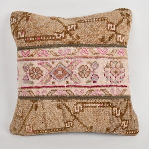 May include: A square decorative pillow with a beige background and a central band featuring floral and geometric patterns in pink, beige, and green. The pillow has a textured, woven appearance, suitable for home decor.