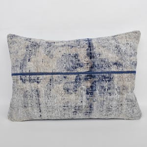 Puede incluir: Cojín decorativo rectangular con un patrón desgastado y texturizado en tonos beige, gris y azul marino. Una franja azul marino recorre horizontalmente el centro, agregando un elemento de contraste.