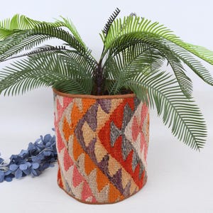 Könnte beinhalten: Ein dekorativer Blumentopf mit einem farbenfrohen geometrischen Muster in Orange-, Rot- und Beigetönen. Der Topf enthält eine grüne Pflanze mit langen, federartigen Blättern. Eine blaue Blumengirlande liegt in der Nähe.