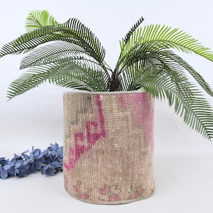 Könnte beinhalten: Ein zylindrischer, beige-rosa gemusterter Pflanztopf mit einer grünen Pflanze im Inneren. Der Pflanztopf hat ein strukturiertes, gewebtes Aussehen. Eine blaue Blumengirlande befindet sich im Vordergrund. Der Pflanztopf ist für den Innen- oder Außenbereich geeignet.