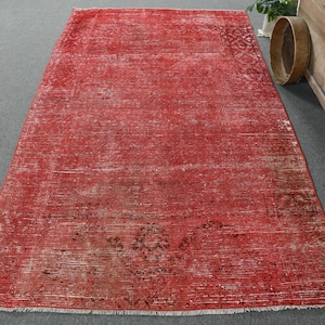 Puede incluir: Una alfombra rectangular roja desgastada, con un aspecto vintage descolorido. La alfombra presenta un campo central rojo con áreas más claras y parches más oscuros, de tono marrón. Los bordes tienen flecos y la alfombra está sobre una superficie gris.