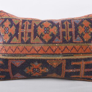 May include: Rectangular decorative pillow with a woven design. The pillow features a pattern of orange, navy blue, and green geometric shapes. The pillow is a home decor item.