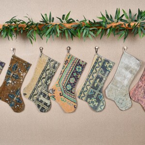 May include: Seven decorative Christmas stockings with patterned designs in various colors, including orange, brown, and blue. The stockings are hanging from small hooks, with a garland of green leaves above.