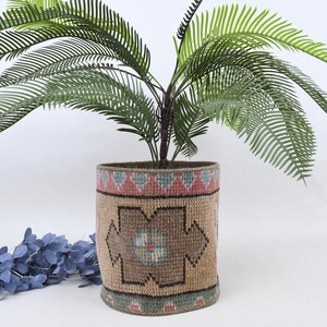Può includere: Un vaso cilindrico tessuto con un motivo geometrico nei toni del marrone, rosso e blu. Il vaso contiene una pianta verde con foglie lunghe e sottili. Una composizione floreale blu poggia sulla superficie.