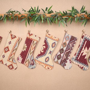May include: Seven Christmas stockings with various geometric patterns in burgundy, cream, and blue hues hang from small hooks. A decorative garland of green leaves and branches sits above the stockings, creating a festive display.