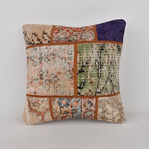 Puede incluir: Una almohada cuadrada de patchwork con una variedad de colores y patrones, incluyendo beige, marrón, verde y morado. La almohada está hecha de una tela suave y tejida y tiene un aspecto único y hecho a mano.