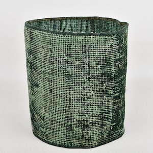 May include: A cylindrical, handmade, green and black rug basket. The basket has a textured, woven appearance. The color is a mix of light green and dark green. The basket is likely used for storage or decoration.
