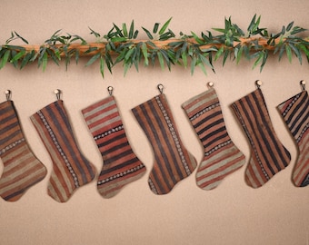 Può includere: Sette calze di Natale con motivi a righe nei toni del marrone, rosso e beige sono appese a piccoli ganci. Una ghirlanda decorativa di foglie e rami verdi è sopra le calze, creando un'atmosfera festosa.