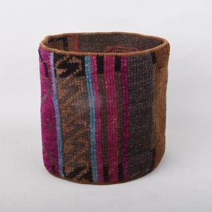 May include: A cylindrical woven basket with a colorful, textured design. The basket features vertical stripes of brown, magenta, and blue, with a dark brown interior. The exterior has a patchwork appearance, with geometric patterns.