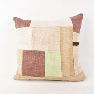 Puede incluir: Cojín decorativo cuadrado con diseño de patchwork. El cojín presenta una mezcla de colores neutros, incluyendo beige, crema, marrón claro y un toque de verde. El cojín está hecho de una tela texturizada y tiene un aspecto suave y acogedor.
