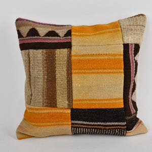 May include: Square patchwork pillow with a variety of woven patterns and colors. The pillow features sections of tan, brown, orange, and black. The design includes stripes, geometric shapes, and a textured appearance, suitable for home decor.