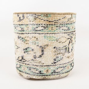 Pode incluir: Um cesto cilíndrico tecido com um design de tapete vintage. O cesto apresenta um fundo creme com padrões em tons de azul, bege e preto. A borda é de cor bege sólida. Este cesto decorativo é adequado para armazenamento.
