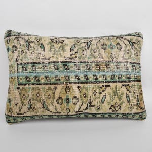 Puede incluir: Cojín decorativo rectangular con diseño de alfombra vintage. El cojín presenta un fondo beige con una banda central de acentos azul claro y verde. El patrón incluye elementos florales y geométricos.