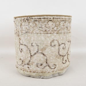 May include: A cylindrical storage basket made from a vintage rug. The basket features a beige and brown pattern with a decorative border. The base is a light gray fabric, and the rim is a darker beige.