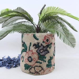 Könnte beinhalten: Ein zylindrischer beigefarbener Pflanztopf mit einem Blumenmuster in Rosa, Grün und Grau, der eine grüne Pflanze mit langen, schlanken Blättern enthält. Blaue Zierblumen befinden sich am Fuß des Pflanztopfs.