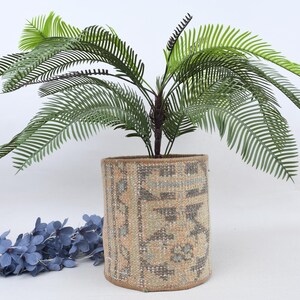 May include: A decorative cylindrical planter with a patterned design, holding a faux plant with green, feathery leaves. The planter is beige and brown, and the plant has a dark brown stem. A blue floral garland is in the foreground.