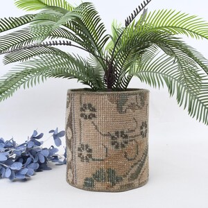 May include: A cylindrical, beige-colored planter with a floral pattern, holding a green plant with long, slender leaves. A blue floral arrangement is in the foreground. The planter has a textured appearance, suggesting a woven or fabric material.