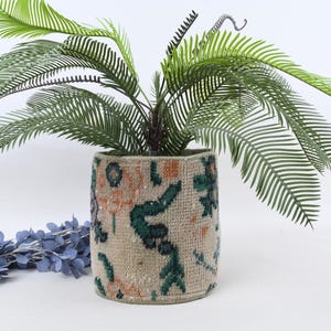 Op de afbeelding: Een cilindrische beige plantenbak met een patroon in groen en oranje, met een levendige groene plant met lange, veervormige bladeren. Een blauw bloemstuk rust aan de linkerkant en creëert een decoratieve weergave.