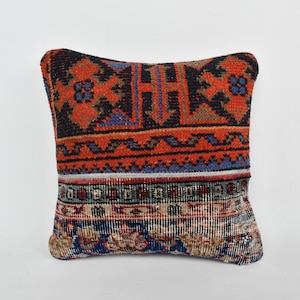 May include: A square pillow with a colorful geometric pattern in shades of red, blue, black, and white. The pillow is made of woven fabric and has a fringed edge.