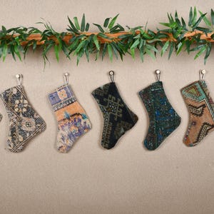 May include: Eight colorful patterned stockings hang from a wooden rod with green leaves. The stockings are made of fabric and have a variety of designs.