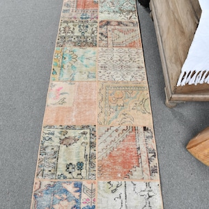 Pode incluir: Um tapete comprido e retangular de patchwork com uma variedade de cores e padrões. O tapete é feito de quadrados menores de tecido, cada um com um design diferente. As cores incluem bege, marrom, azul, rosa e verde.