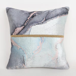 May include: Square decorative pillow with a marbled design in shades of gray, blue, and pink. A band of tan fabric separates the top and bottom sections. The pillow is a home decor item.