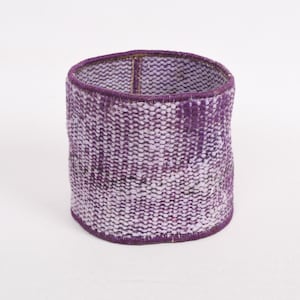 Puede incluir: Una cesta cilíndrica tejida en tonos morados y blancos. La cesta tiene una parte superior redondeada y un borde morado oscuro. El patrón de tejido es visible, creando una apariencia texturizada. La cesta está sobre un fondo blanco.
