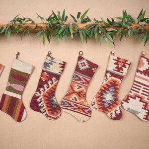 May include: Seven Christmas stockings with geometric patterns in red, orange, and cream hang from hooks. A garland of green leaves and branches is above the stockings, creating a festive holiday display.