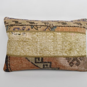 Puede incluir: Cojín decorativo rectangular con diseño patchwork. El cojín presenta una mezcla de colores, incluyendo beige, naranja, marrón y verde. El cojín tiene un aspecto texturizado, lo que sugiere una construcción tejida o de tela. El diseño incluye patrones geométricos y abstractos.