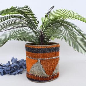 Puede incluir: Una maceta tejida con un diseño geométrico en naranja, gris y negro contiene una planta verde con hojas largas y plumosas. La maceta es cilíndrica y se encuentra sobre una superficie blanca. Hay tallos decorativos azules en primer plano.