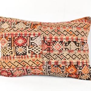 May include: Rectangular decorative pillow with a patchwork design. The pillow features a mix of geometric patterns in shades of beige, orange, red, and brown. The pillow is likely made of a woven fabric and is placed against a white background.