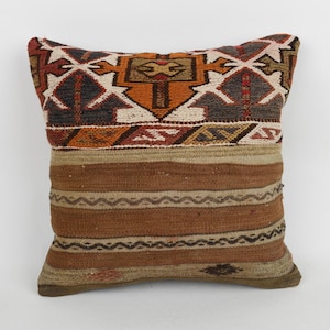 Op de afbeelding: Vierkant decoratief kussen met een geweven ontwerp. Het bovenste gedeelte heeft een geometrisch patroon in tinten rood, oranje, bruin en wit. Het onderste gedeelte heeft horizontale strepen in bruin en beige. Het kussen is vierkant.