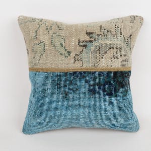 May include: Square decorative pillow with a two-tone design. The top half is a beige color with faded patterns, and the bottom half is a textured teal blue. A thin brown band separates the two sections. The pillow is likely made of fabric.