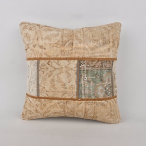 Op de afbeelding: Vierkant beige kussen met een patchwork ontwerp. Het heeft een centrale band van stof met patronen in groen, beige en grijs, omlijst door een bruine rand. De hoofdgedeelte van het kussen heeft een textuur, bloemenpatroon.