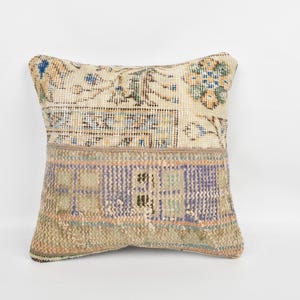 Pode incluir: Almofada decorativa quadrada com design patchwork. A almofada apresenta uma parte superior creme com padrões florais azuis e verdes e uma seção inferior com um padrão geométrico roxo e bege. A almofada está em um fundo branco.