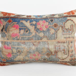May include: Rectangular pillow with a vintage, distressed look. The pillow features a woven design with a central band and floral patterns in pink, blue, and orange hues. The pillow's edges are trimmed with a darker border.