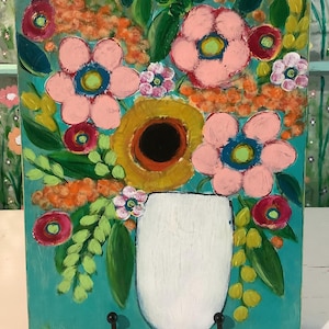 May include: A rectangular painting on a teal background featuring a white vase filled with a bouquet of flowers. The arrangement includes large pink blossoms with blue centers, a central golden-yellow flower, and various green leaves and orange accents.
