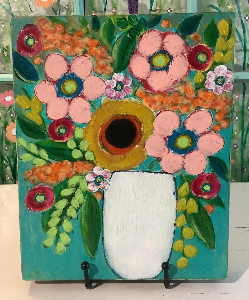 May include: A rectangular painting on a teal background featuring a white vase filled with a bouquet of flowers. The arrangement includes large pink blossoms with blue centers, a central golden-yellow flower, and various green leaves and orange accents.