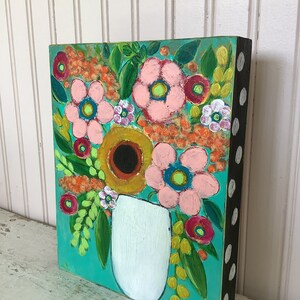 May include: A rectangular painting on a wooden block featuring a white vase filled with a vibrant floral arrangement. The flowers are painted in shades of pink, yellow, and orange against a bright teal background. The side of the block is black with white dots.