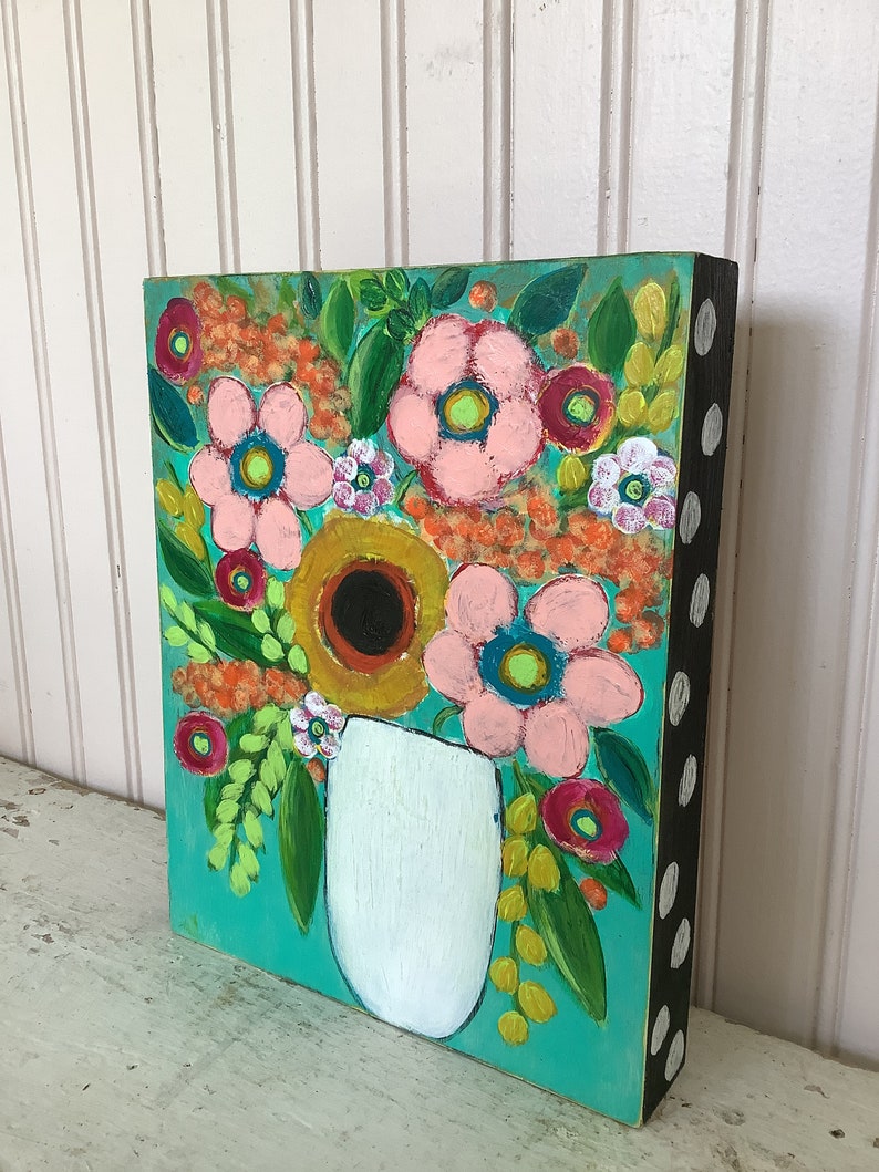 May include: A rectangular painting on a wooden block featuring a white vase filled with a vibrant floral arrangement. The flowers are painted in shades of pink, yellow, and orange against a bright teal background. The side of the block is black with white dots.