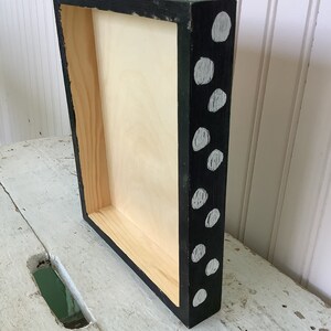 May include: A rectangular wooden box frame with a natural light-colored interior. The exterior sides are painted black and feature a pattern of irregular, light gray circular spots distributed across the surface of the frame edges.