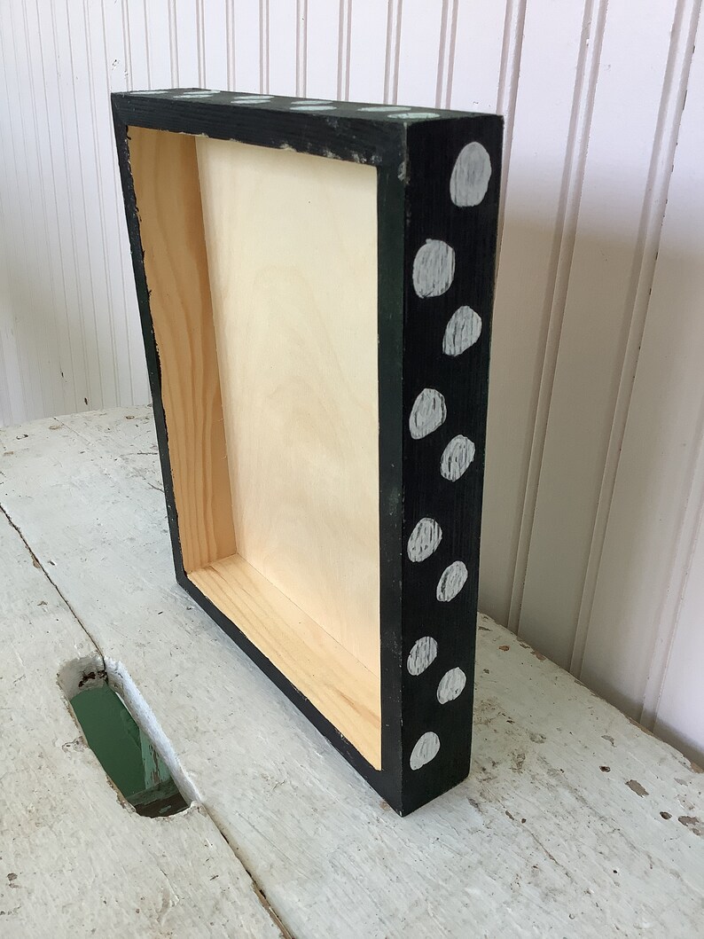 May include: A rectangular wooden box frame with a natural light-colored interior. The exterior sides are painted black and feature a pattern of irregular, light gray circular spots distributed across the surface of the frame edges.