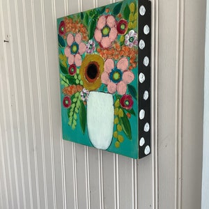 May include: A square canvas painting featuring a white vase filled with a colorful floral arrangement on a teal background. The side of the canvas is painted black with white polka dots, showcasing a variety of pink, orange, and red flowers.