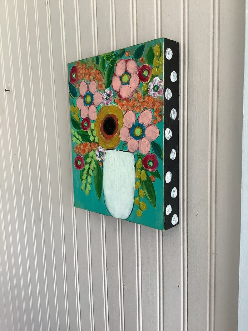 May include: A square canvas painting featuring a white vase filled with a colorful floral arrangement on a teal background. The side of the canvas is painted black with white polka dots, showcasing a variety of pink, orange, and red flowers.