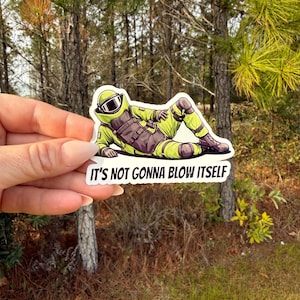 May include: A sticker featuring a cartoon illustration of a person in a bomb suit, lying down. The person is wearing a green suit, helmet, and goggles. The sticker has the text "IT'S NOT GONNA BLOW ITSELF".